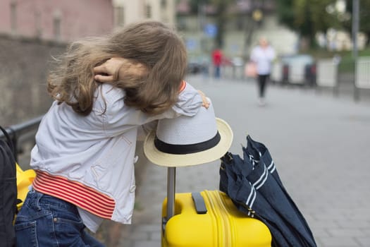 Concept of tourism, travel, little girl with luggage suitcase, backpack, hat, umbrella, close-up in tourist city