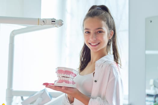 Smiling teenager girl in dental office holding jaw model with teeth in her hands