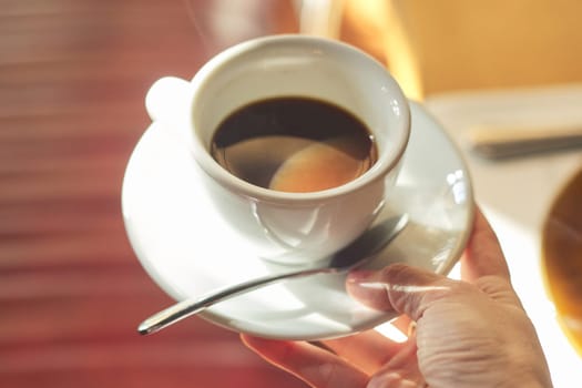 Cup with morning coffee in female hand, cap in white cup with saucer spoon closeup, lighting morning sun