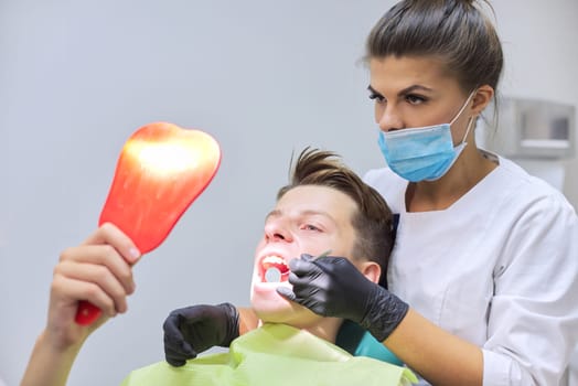 Female dentist doctor treats teeth to teenage boy sitting in dental chair looking at teeth in mirror