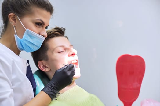 Female dentist doctor treats teeth to teenage boy sitting in dental chair looking at teeth in mirror