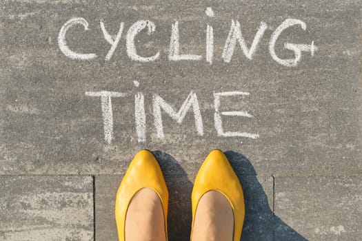 Cycling time, text on gray sidewalk with woman legs, top view.