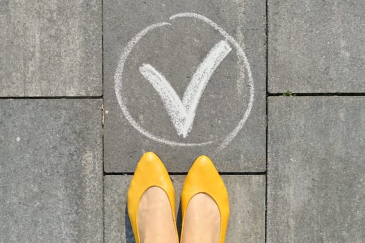 Checkmark ok sign on gray sidewalk with woman legs, top view.