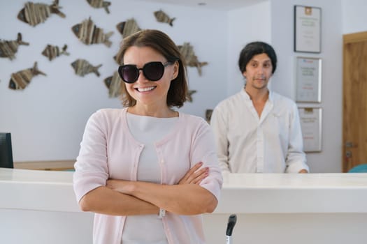 Smiling happy woman guest in lobby interior of resort spa hotel, male receptionist working near reception desk