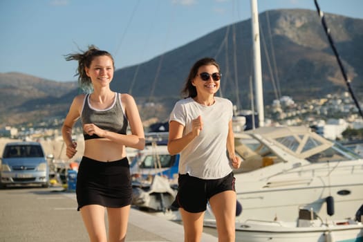 Smiling mother and daughter teenager jogging running at seaside promenade together, sports outdoor, background sea mountains city, sunny summer day