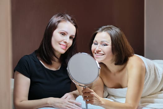 Talking and smiling women beautician and mature female patient with mirror in beauty salon. People, spa, cosmetology and skincare concept