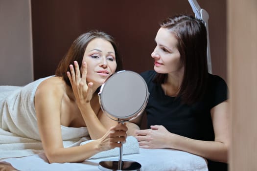 Talking and smiling women beautician and mature female patient with mirror in beauty salon. People, spa, cosmetology and skincare concept