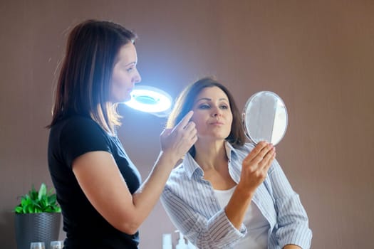 Middle-aged woman in beauty salon, female talking to a beautician and looking at her face skin hair in the mirror. People, spa, cosmetology and skincare concept