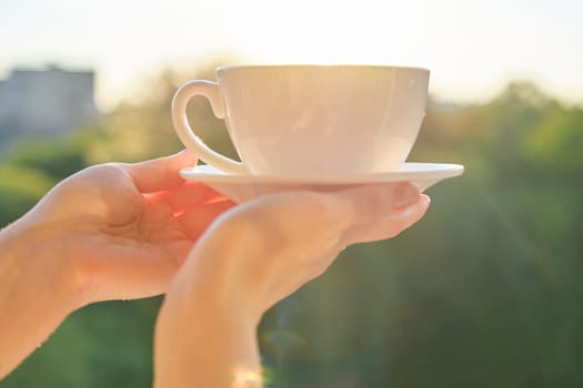 Set of white ceramic cup with saucer in womans hand, background sunset, evening city