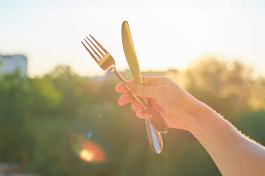 Steel metal shiny knife and fork in a womans hand, background sunset, evening city