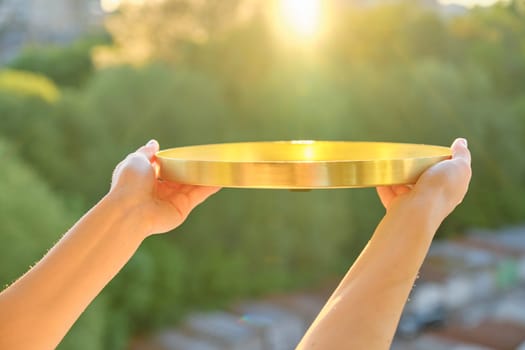 Round golden brass metal tray in womans hands, background sunset, evening city
