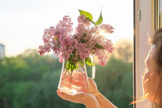 Bouquet of flowers lilac in jug in womans hand, background sunset, evening city