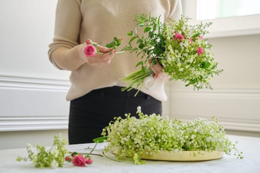 Woman florist with different flowers on the table making bouquet, spring flowers of lily of the valley, roses, viburnum. Floristry, work of florist, flower shop, beauty