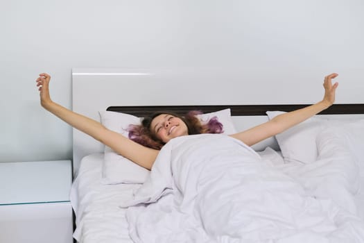 Teen girl waking up in the morning lying on pillow in bed with arms open.
