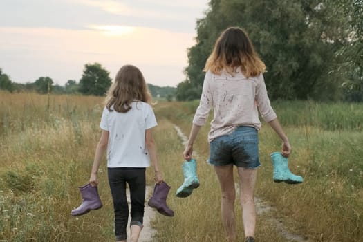 Two happy girls sisters walking after rain in dirty clothes with rain boots in hands, back view, rural road summer nature background