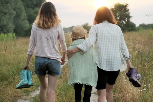Family mother and two daughters walking together along country road after summer rain holding rubber boots in hands, back view, nature background golden hour
