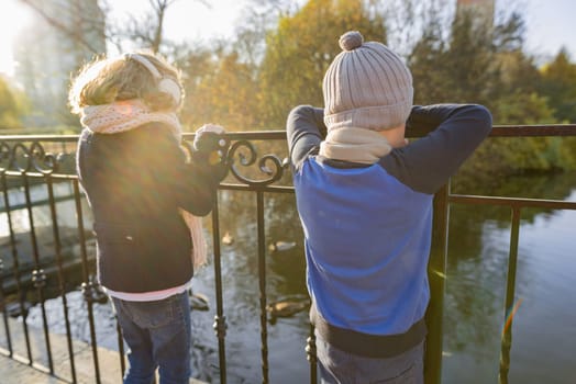 Children boy and girl standing backs on bridge, looking at ducks, sunny autumn day in park, golden hour