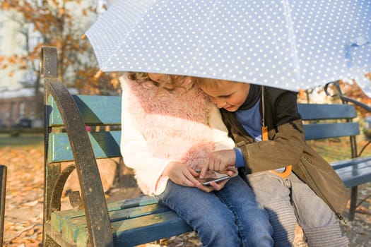 Outdoor portrait of two smiling children boy and girl, sitting under an umbrella on bench in autumn park, using smartphone, golden hour