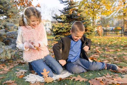 Happy kids boy and girl playing and sitting on yellow leaves in sunny autumn park, leaf fall golden hour