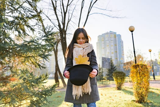 Autumn outdoor portrait of girl with yellow maple leaves, park background at golden hour, child in coat hat