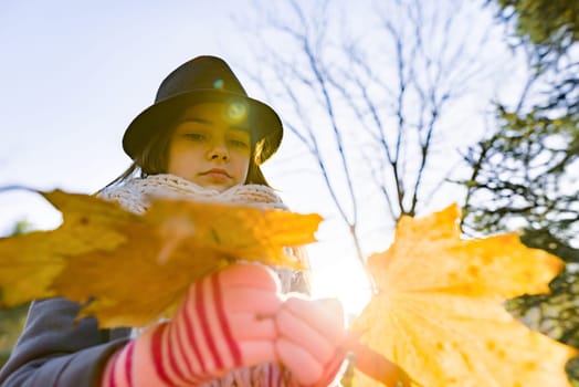 Autumn outdoor portrait of girl with yellow maple leaves, park background at golden hour, child in coat hat