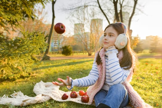 Child girl playing with red apples, sitting in sunny autumn park, healthy food in nature, leaf fall golden hour