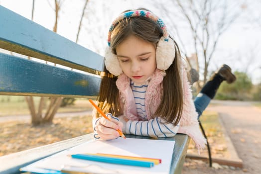 Little beautiful artist drawing with colored pencils, girl sitting on a bench in sunny autumn park, golden hour