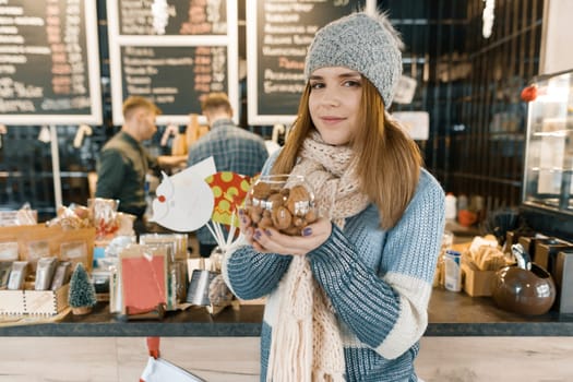 Winter portrait of young beautiful woman in knitted scarf, knitted hat, mittens, warm sweater with bowl of hazelnuts and walnuts. Girl near bar counter, Christmas decorated tree in coffee shop.