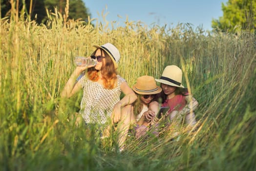 Portrait of three girls sitting and resting in grass, children talking, looking at smartphone laughing on sunny summer day, sunset in meadow