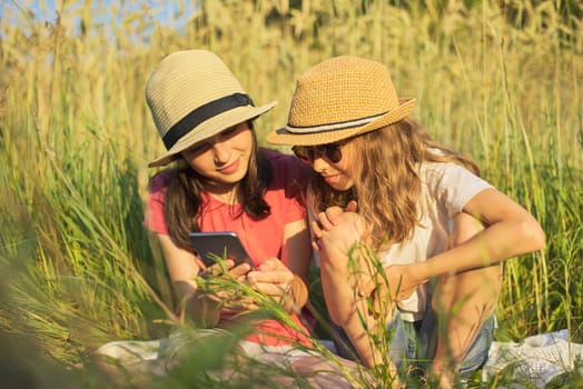 Summer portrait of two girls sitting in grass playing and looking at smartphone, beautiful children at sunset