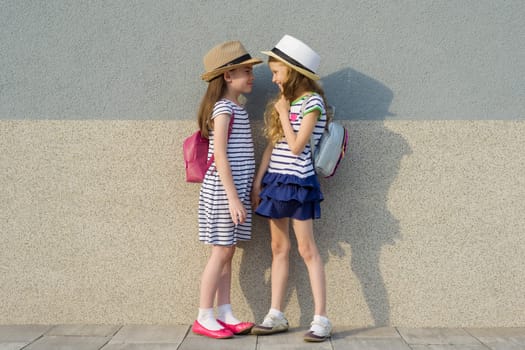 Outdoor summer portrait of two happy girl friends 7,8 years in profile talking and laughing. Girls in striped dresses, hats with backpack, background gray wall