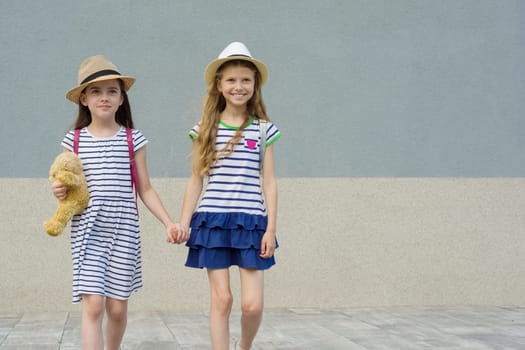 Two little beautiful girlfriends holding hands, girls walking in striped dresses, hats with backpack, background gray wall, copy space