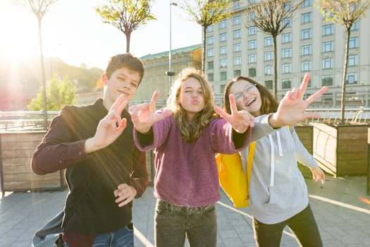 Portrait of friends teen boy and two girls smiling, making funny faces, showing victory sign in the street. City background, golden hour