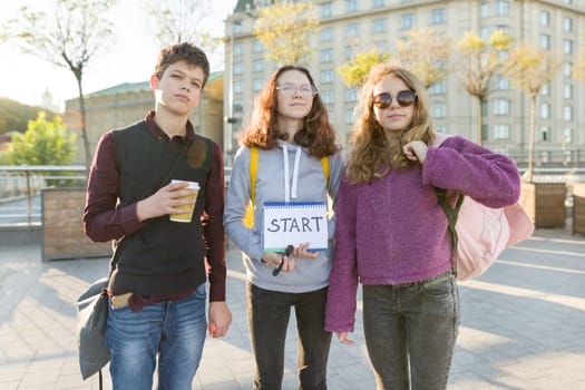 Group teenagers boy and two girls, with a notepad with handwritten word start. Teenagers looking forward, city background, golden hour