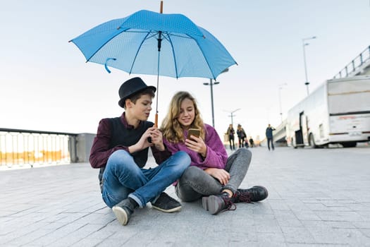 Couple of smiling teen friends sitting under an umbrella and looking at smartphone, lifestyle of teenagers in the city