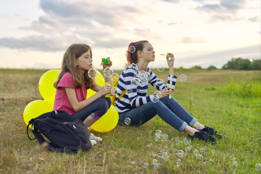 Two girls sister resting in nature, summer vacation in evening near the lake on lawn, children with soap bubbles