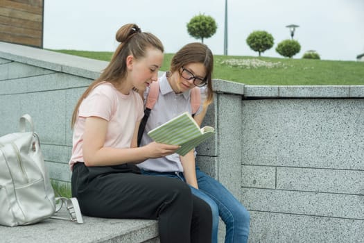 Outdoor portrait of two young beautiful girls students with backpacks, books. Girls talking, looking in book, urban background