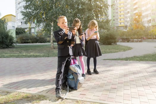 Children of elementary age with smartphones, backpacks, outdoor background. Education, friendship, technology and people concept