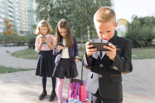 Children of elementary age with smartphones, backpacks, outdoor background. Education, friendship, technology and people concept