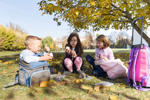 Autumn portrait of children with lunch boxes, school backpacks. Cheerful schoolchildren eat fruit, laughing, talking. Healthy food and healthy lifestyle