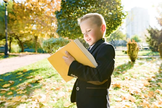 Outdoor autumn portrait of schoolboy reading book, background of yellow trees in the park, boy in jacket, golden hour, back to school