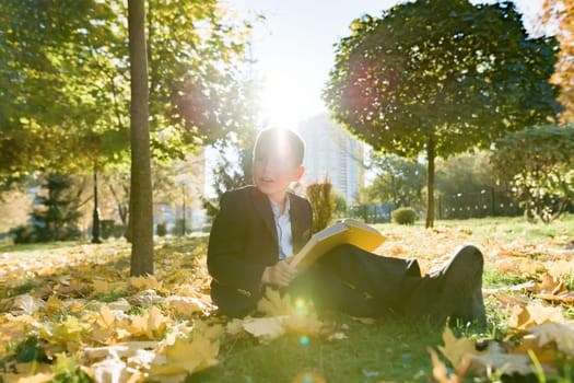 Outdoor autumn portrait of schoolboy reading book, background of yellow trees in the park, boy in jacket, golden hour, back to school