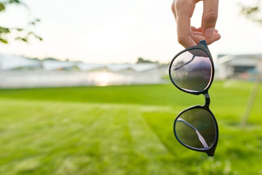 Sunglasses in woman hand close up, copy space, summer green grass background, sunset sky