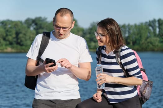 Outdoor mature couple using smartphone, man and woman talking walking in the park, people in sportswear with bottle of water, summer sunny evening near the river