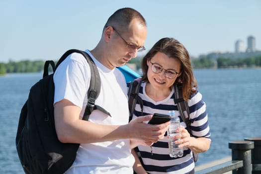 Outdoor mature couple using smartphone, man and woman talking walking in the park, people in sportswear with bottle of water, summer sunny evening near the river