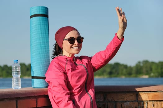 Middle-aged sports woman with yoga mat and bottle of water, an outdoor evening sunny park near the river. Active healthy lifestyle of an age female