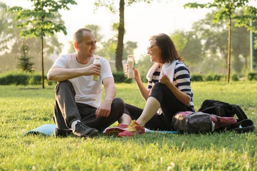 Happy mature couple sitting in the park on fitness mat, resting drinking yogurt after sports exercises. Healthy lifestyle and food age people