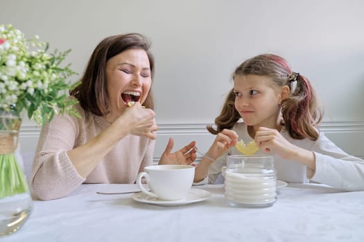 Laughing mother and little daughter, sitting at home at the table drinking from cups and eating lemon. Parent and child having fun and talking