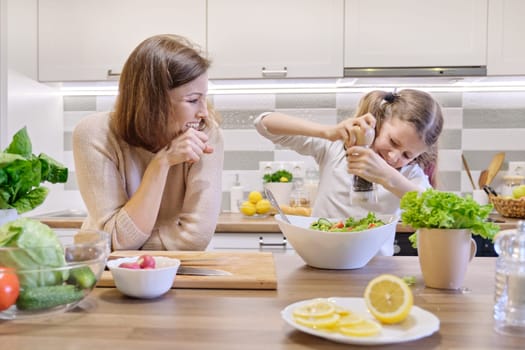 Cooking healthy home meal by family. Mother and daughter cut vegetables at home in the kitchen for salad. Girl salts freshly cooked salad, mother looks up and rejoices