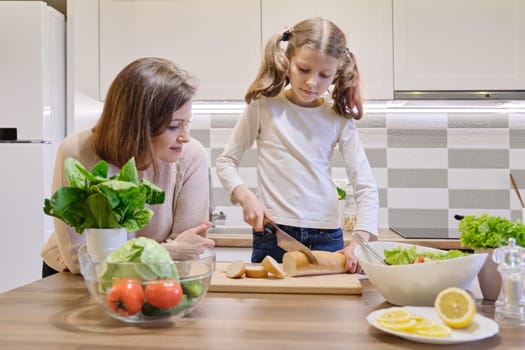 Mother and daughter cooking together in the kitchen at home, girl cutting bread.